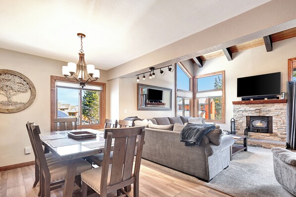 A spacious living area with a fireplace, a dining table with chairs, a sofa, and a television mounted on the wall.