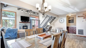 Ein geräumiges Wohnzimmer mit Kamin, einer Essecke mit Holztisch und Stühlen, einem großen Fernseher und einer Treppe, die eine weitere Ebene hinaufführt.