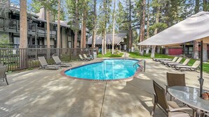 A pool area with lounge chairs and a table, surrounded by tall trees and a building with balconies. - Sunshine Village 157: Pet Friendly, Centrally Loca (Mammoth Lakes)
