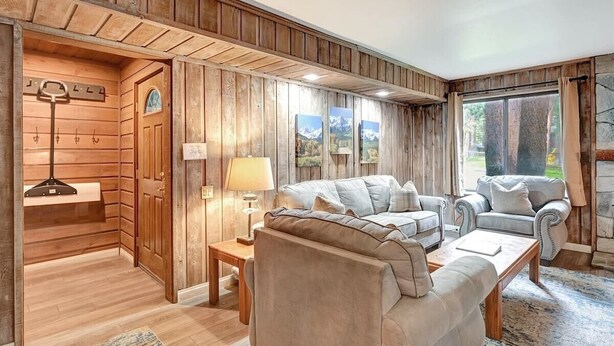 A cozy living room with wooden paneling, a sofa, a chair, and a coffee table. There is a door with a coat rack and a lamp on a side table. - Sunshine Village 157: Pet Friendly, Centrally Loca (Mammoth Lakes)