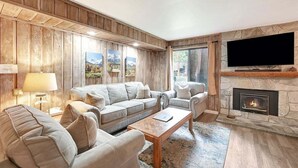 A cozy living room with a stone fireplace, a large flat-screen TV mounted above it, and a wooden paneled wall. - Sunshine Village 157: Pet Friendly, Centrally Loca (Mammoth Lakes)