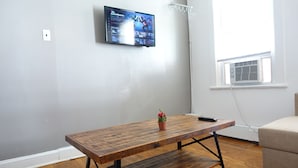 Una habitación con una mesa de madera, una planta en maceta, un televisor en la pared y una ventana con un equipo de aire acondicionado.