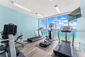 Une salle de sport équipée de tapis de course, de vélos elliptiques et d’un vélo d’appartement, avec une grande fenêtre donnant sur l’océan.
