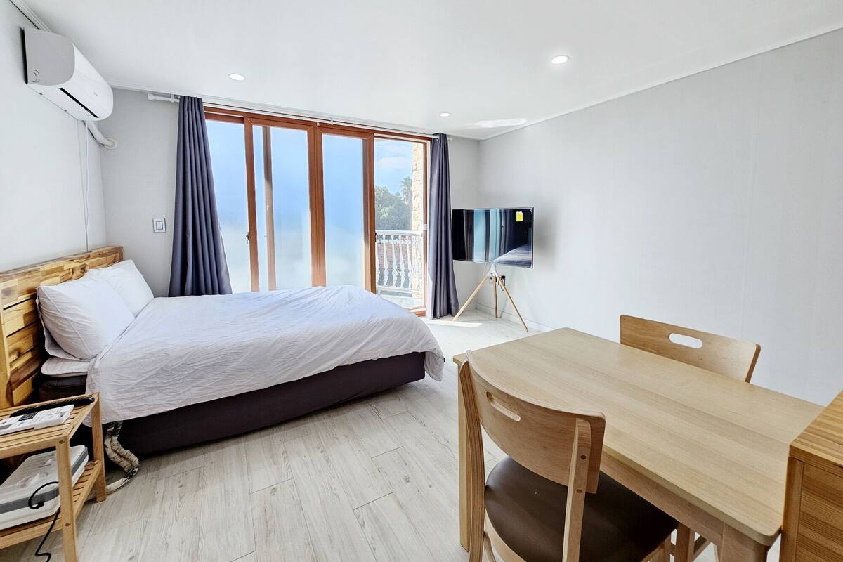 A modern hotel room with a bed, a desk, a chair, a TV on a stand, and a balcony with curtains.