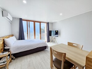 A modern hotel room with a bed, a desk, a chair, a TV on a stand, and a balcony with curtains. - Green Side Pension (Seogwipo)