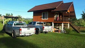 A two-story house with a brown roof and a balcony. There are two pickup trucks parked in front. - Rai Monsicha Khaokho (Khao Kho)