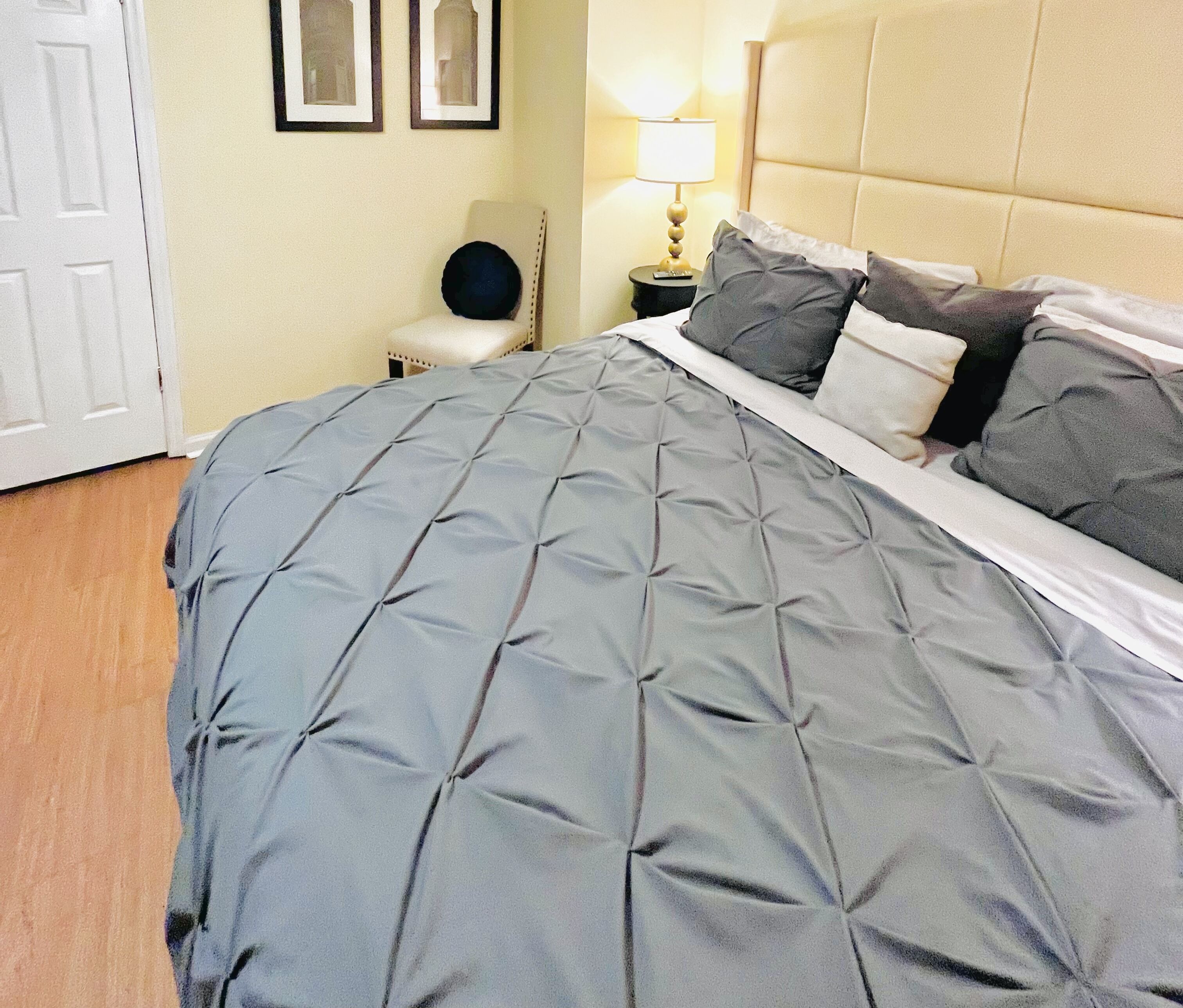 Un lit bien fait, recouvert d’une couverture grise, une lampe de chevet et une chaise placée contre un mur orné de tableaux encadrés.