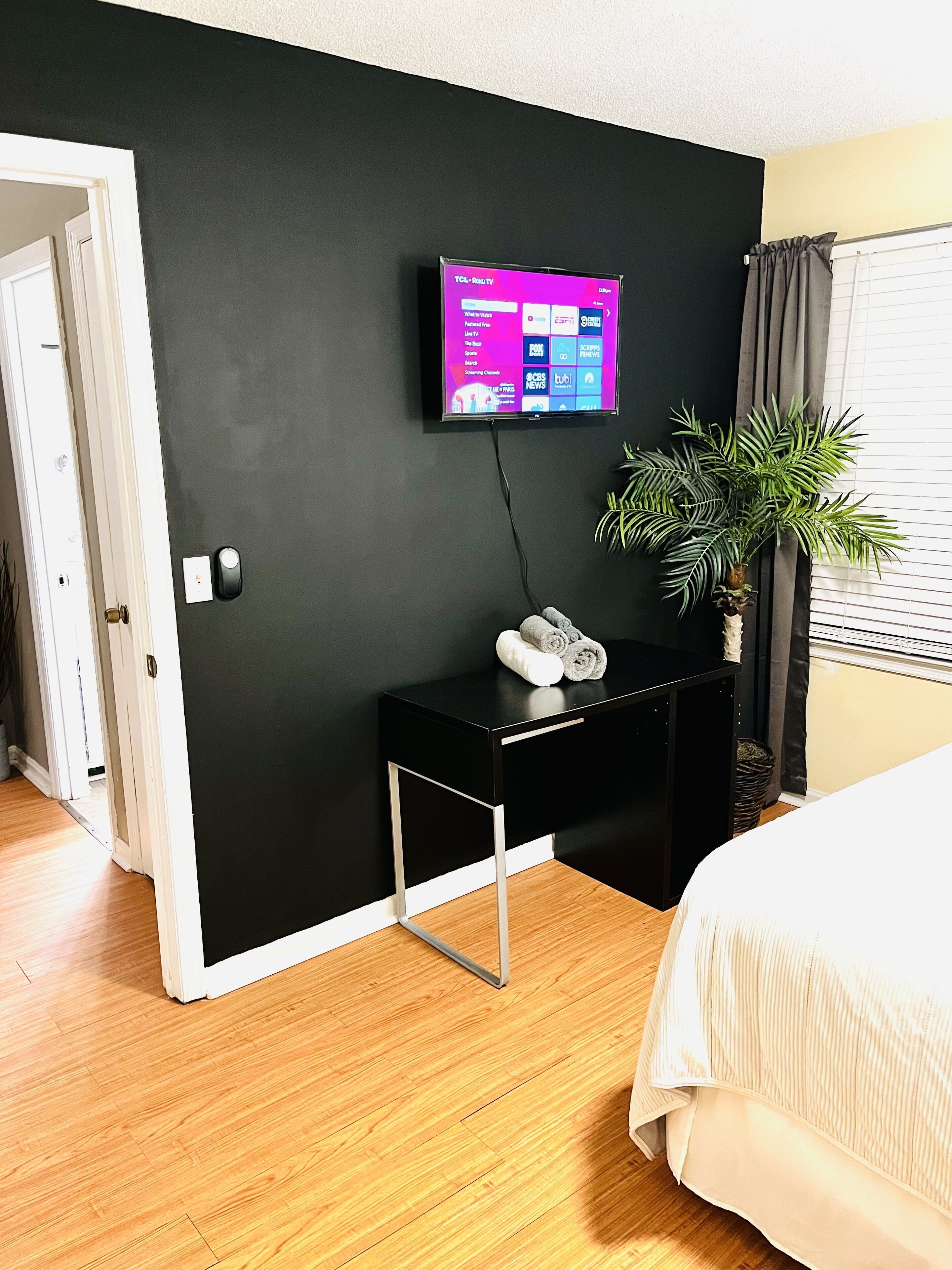 Quarto de hotel com parede preta, televisão montada, mesinha lateral preta pequena com uma planta e uma cama com colcha branca.