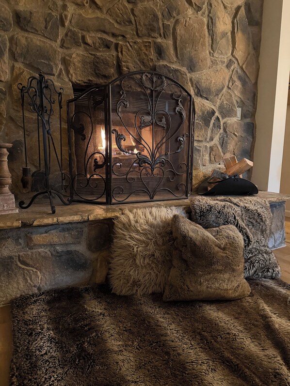A stone fireplace with a wrought iron screen, a log fire, and a fur throw on a wooden bench.