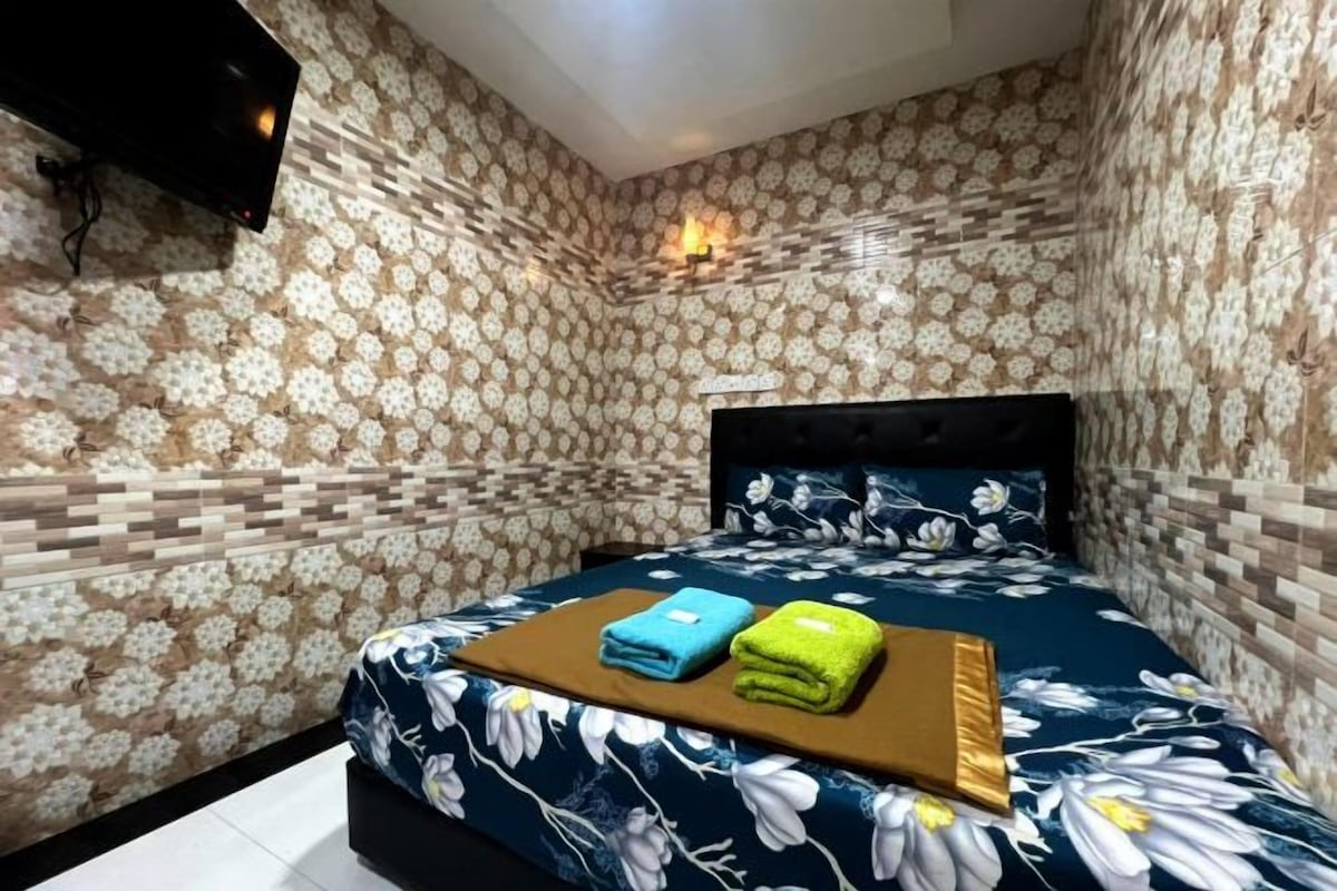 Un dormitorio con pared de estampado floral, una cama con colcha azul floral y dos toallas sobre una mesita de noche.