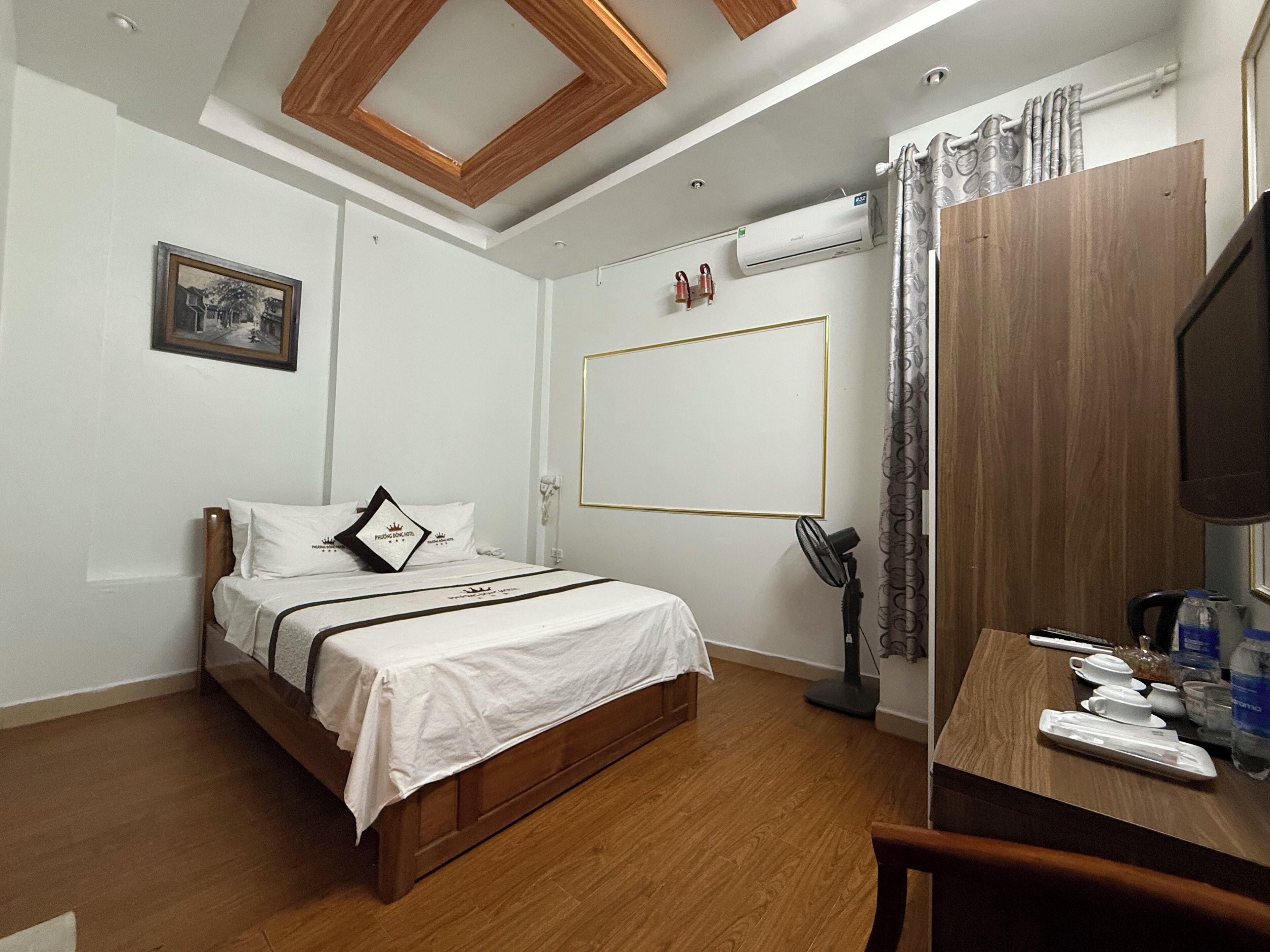 A hotel room with a bed, a desk with a chair, a flat-screen TV, an air conditioning unit, and a window with curtains.