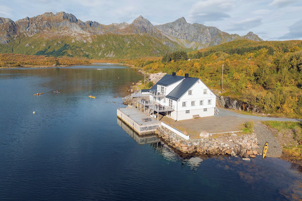 A white building with a dock extending into a lake, surrounded by mountains and autumn foliage.