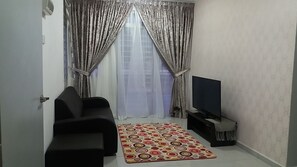 Un salón con un sofá negro, un televisor sobre un mueble bajo, un tapete estampado y cortinas de gasa.