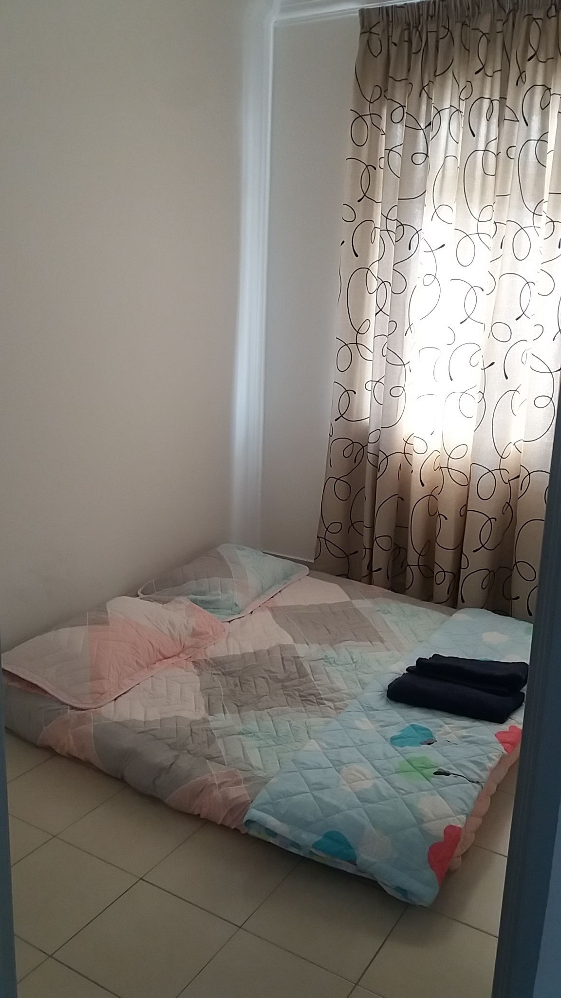 Een klein, verzorgd bed met een gewatteerde spreideken en kussens, een raam met gebloemde gordijnen en een betegelde vloer.