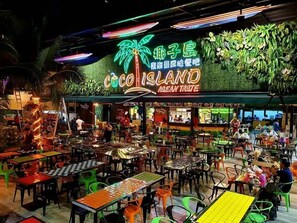 A vibrant indoor dining area with multiple tables and chairs, a neon sign reading "COCO ISLAND," and a green palm tree decoration. - Wangs Hotel Gurney Drive (George Town)