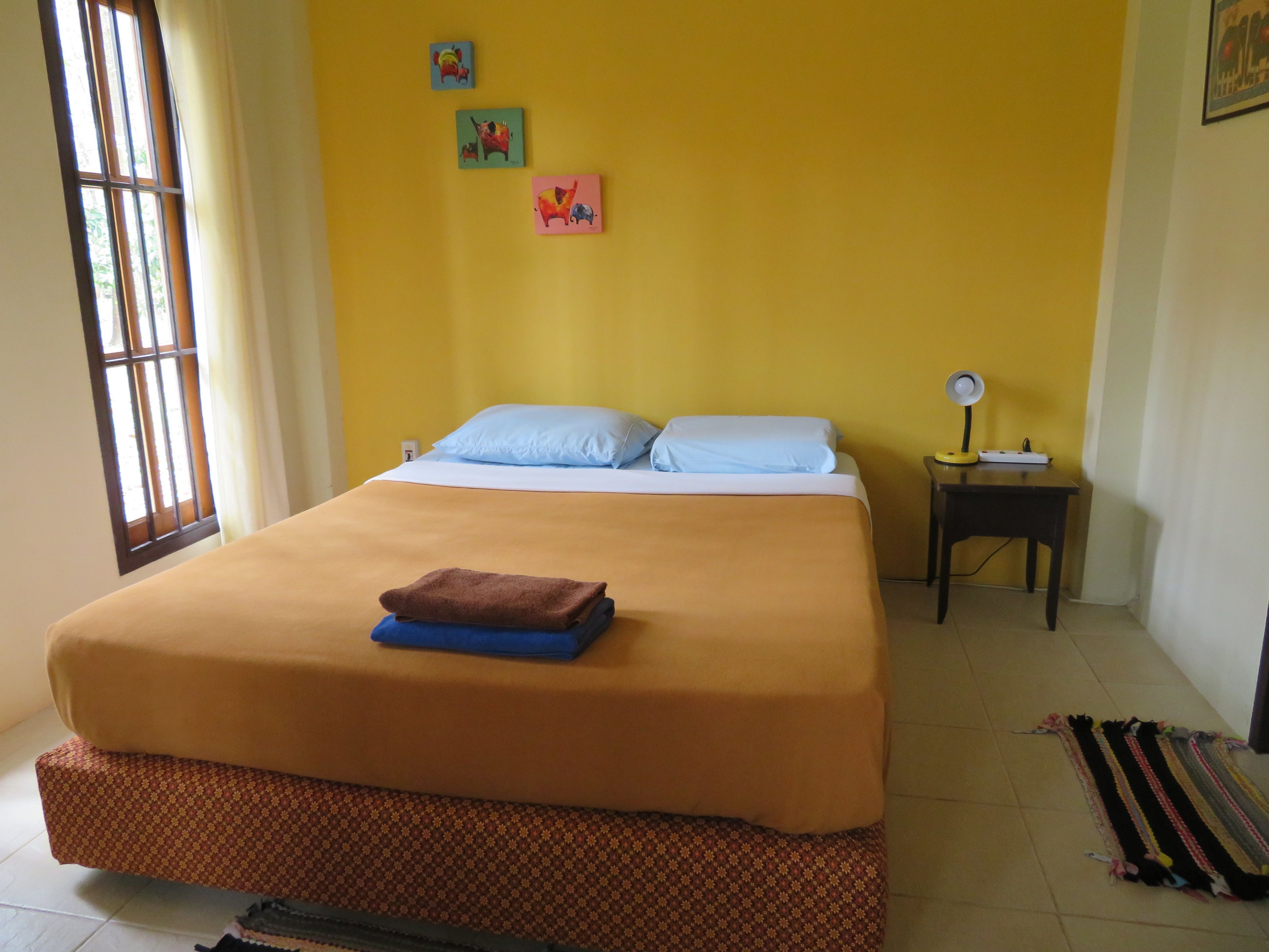 A bed with a brown bedspread and two pillows, a small bedside table with a lamp, and a window with curtains.