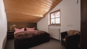 Quarto com teto inclinado, janela com vista, radiador, cama, cadeira e mesa de cabeceira com abajur.