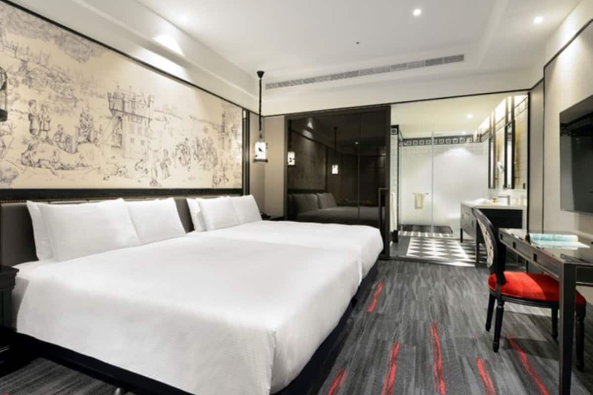 A hotel room with a large bed, a desk, and a chair. There is a large window and a painting on the wall.