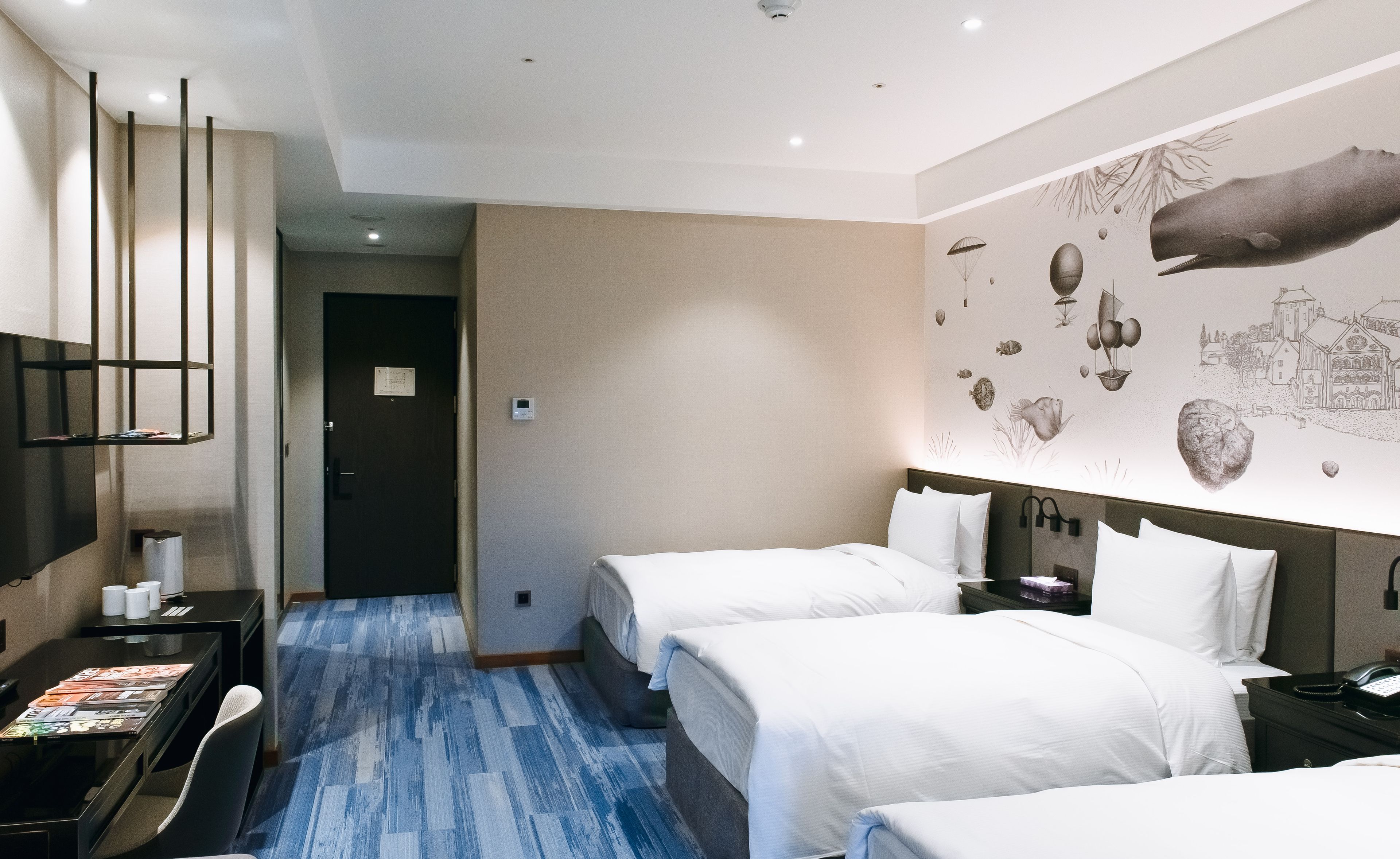 Ein Hotelzimmer mit zwei Betten, einem Schreibtisch mit Stuhl, einem Flachbildfernseher und Wandgemälde.