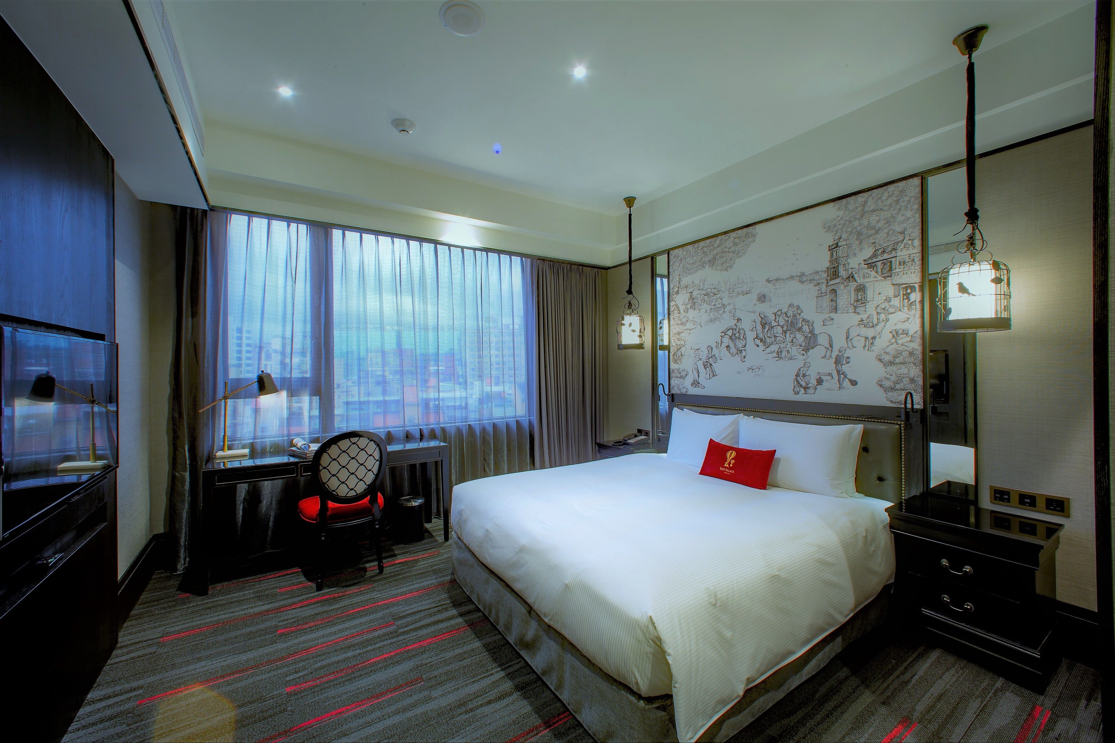 A hotel room with a large bed, a desk with a chair, a television, and a view of the city through the window.