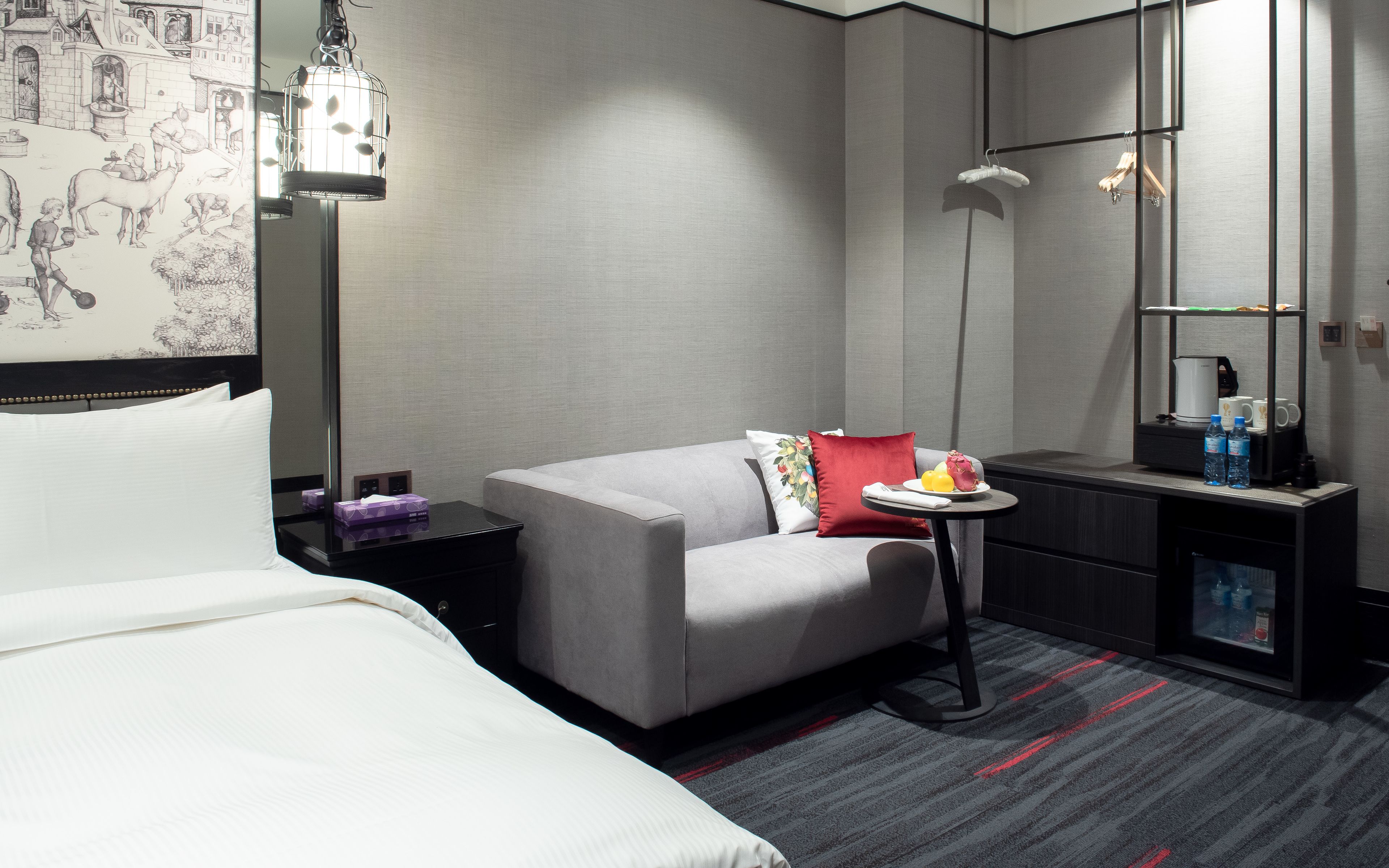 A modern hotel room with a bed, a grey sofa, a small table with a fruit plate, a coffee machine, and a minibar.