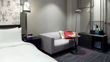 A modern hotel room with a bed, a grey sofa, a small table with a fruit plate, a coffee machine, and a minibar.