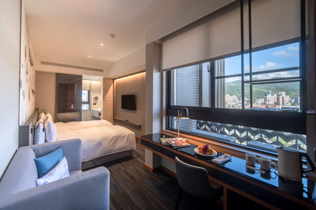 A modern hotel room with a bed, a desk with a computer, a chair, a sofa, a TV, and a large window with a city view.