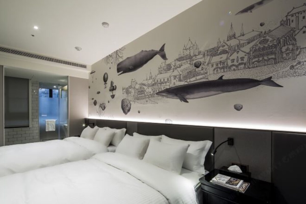 A hotel room with a large bed, a nightstand, a wall mural of marine life and a cityscape, and a modern bathroom visible through an open door.