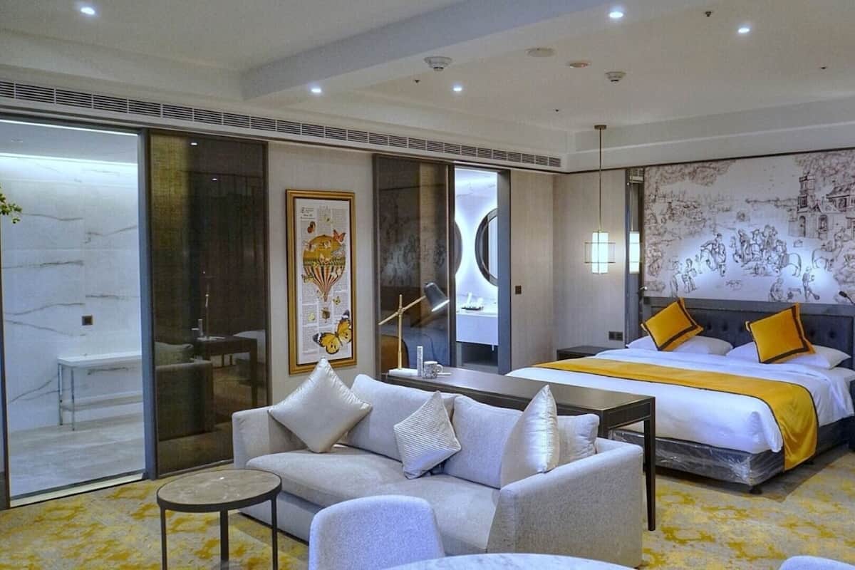 A modern hotel room with a large bed, a sofa, a small table, and a large window.