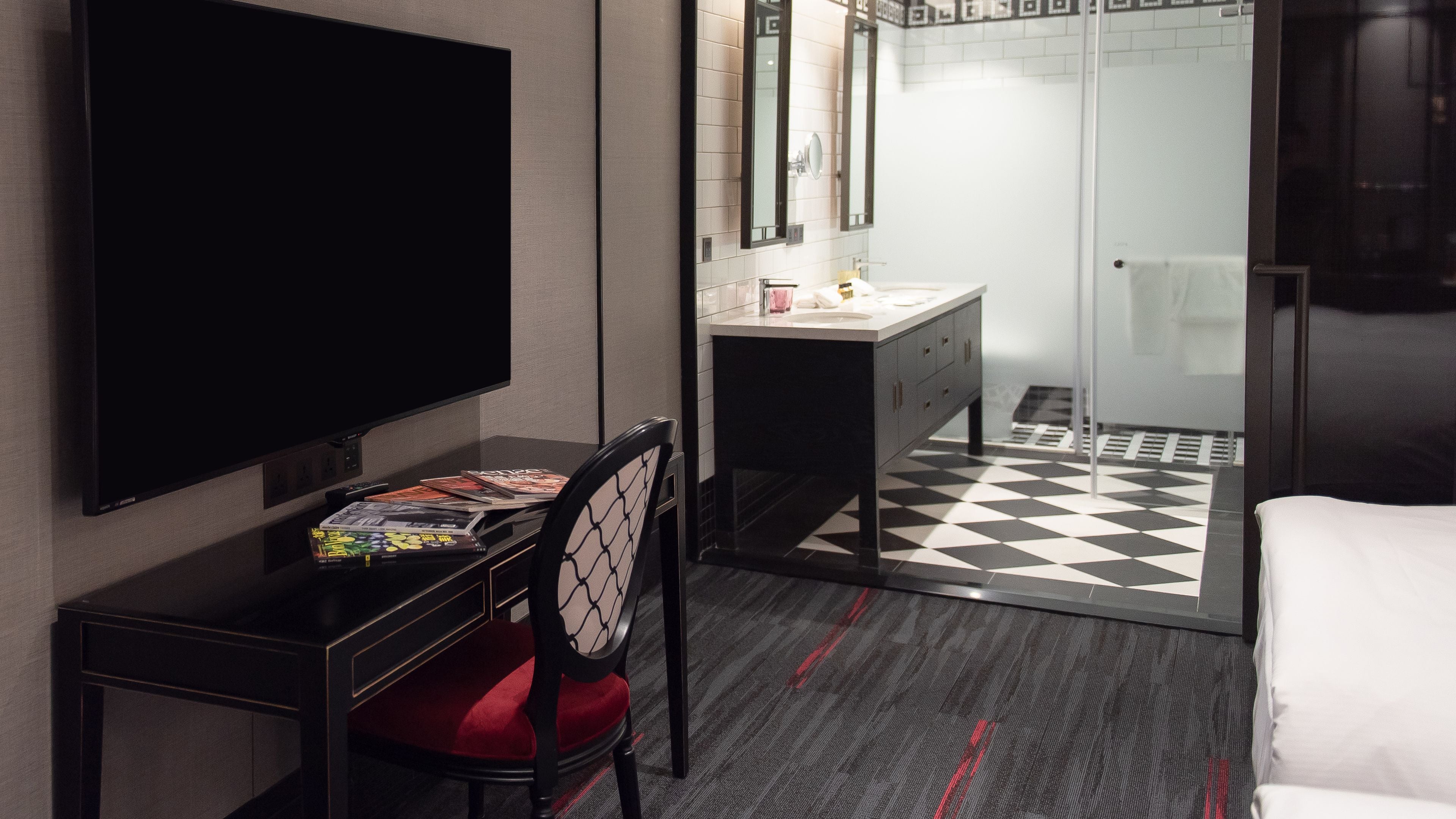 A hotel room with a flat-screen TV, a desk with a chair, a vanity with a mirror, a bed, and a checkered floor.