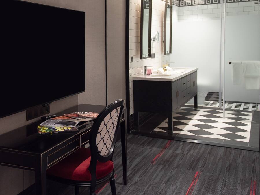 Ett hotellrum med en platt-TV, ett skrivbord med en stol, en sminkbord med en spegel, en säng och ett schackrutigt golv.