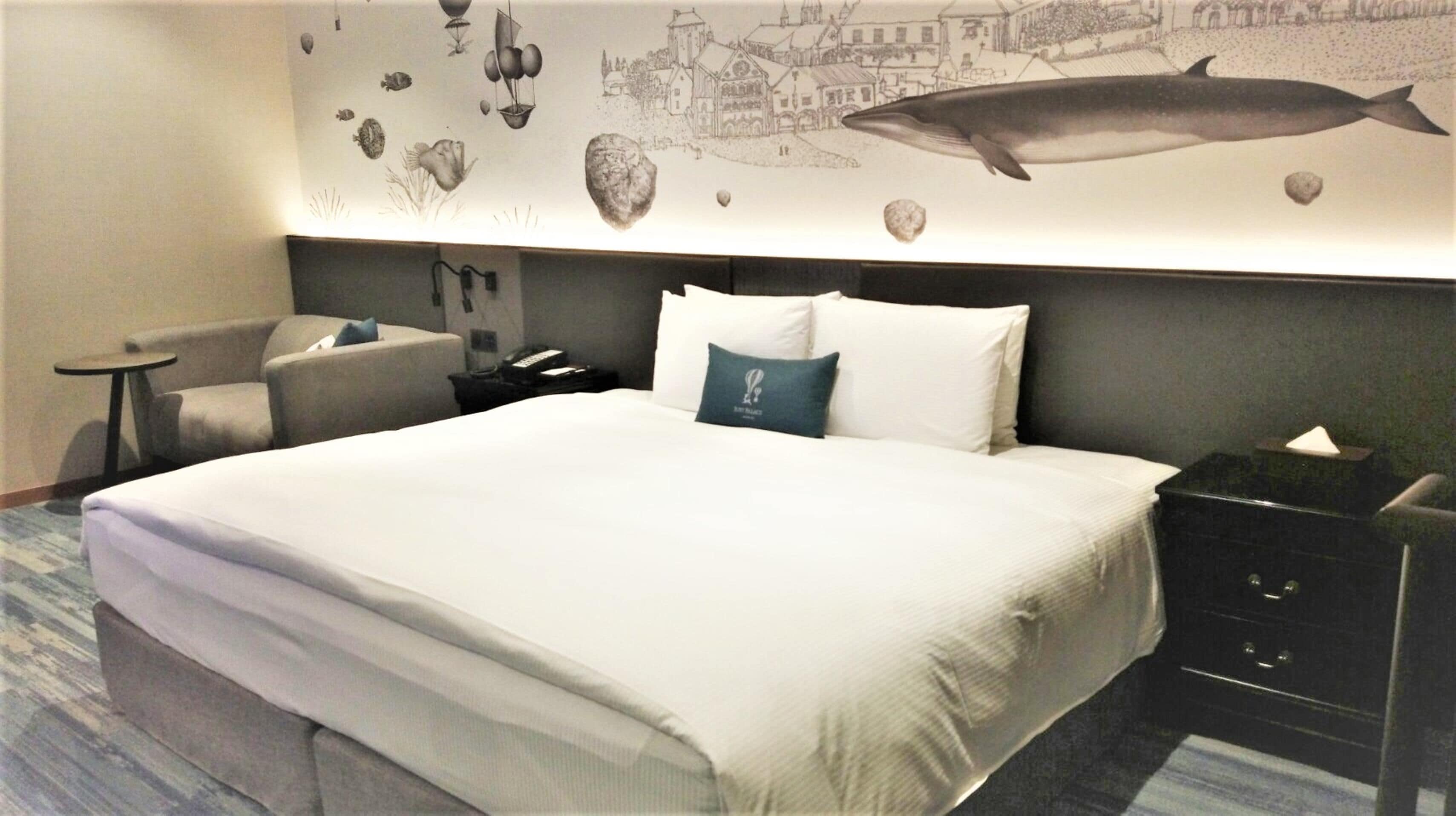 A hotel room with a large bed, a desk, a chair, and a wall mural depicting marine life and a town scene.