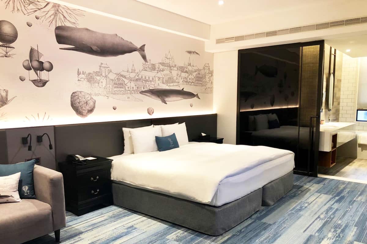 A modern hotel room with a large bed, a nightstand, a chair, and a wall mural.