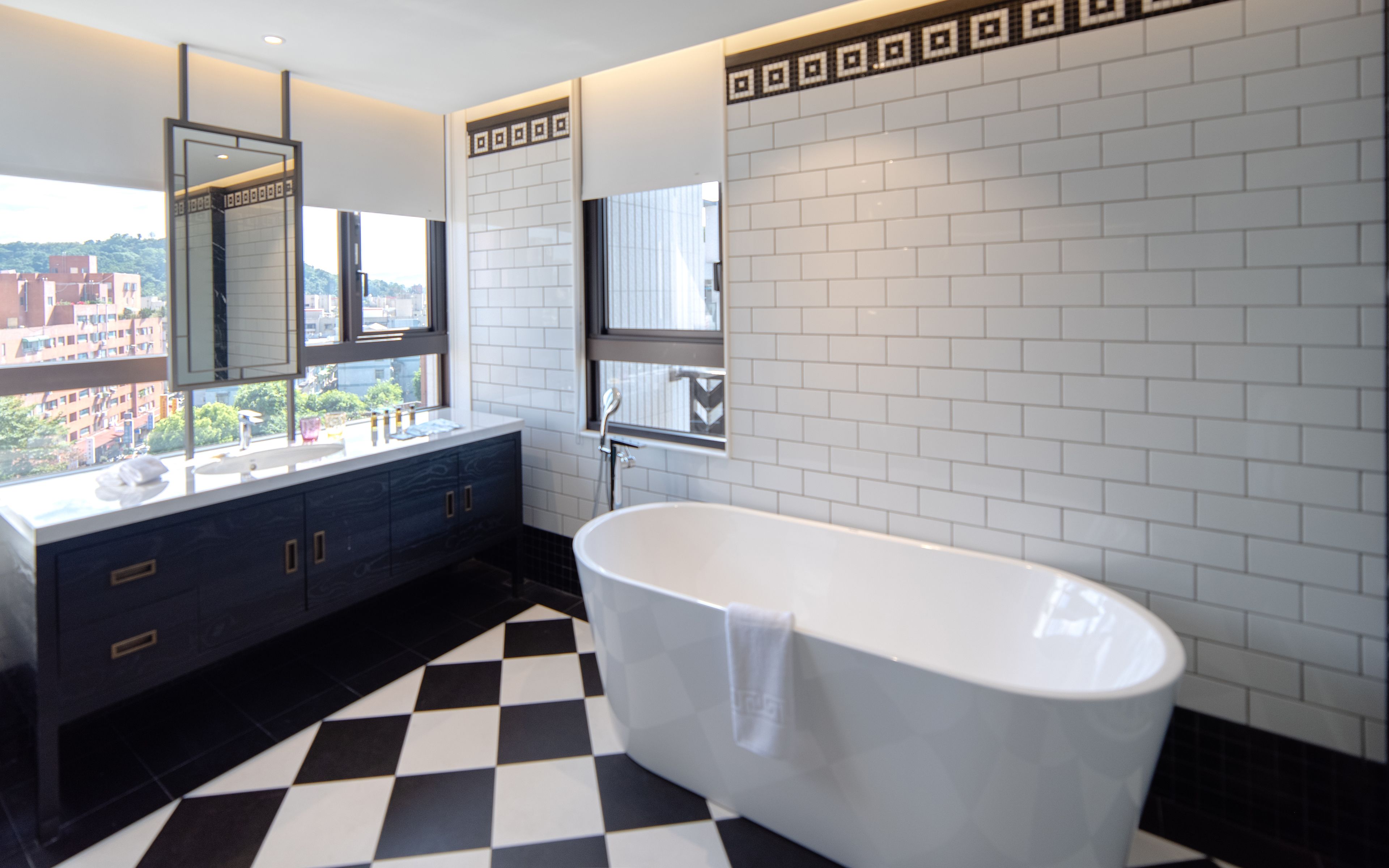 A modern bathroom with a checkered floor, a freestanding bathtub, and a large window offering a city view.
