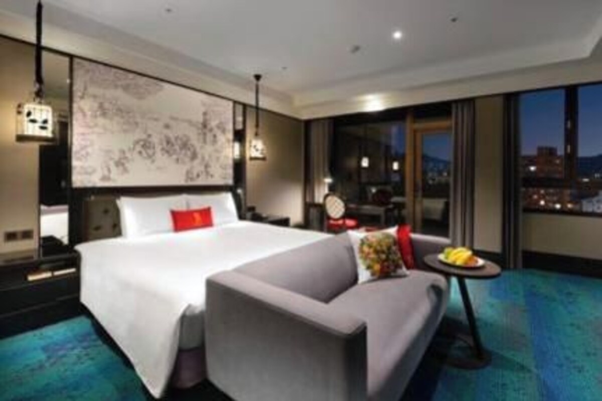 A hotel room with a large bed, a sofa, a small table with fruit, and a city view.