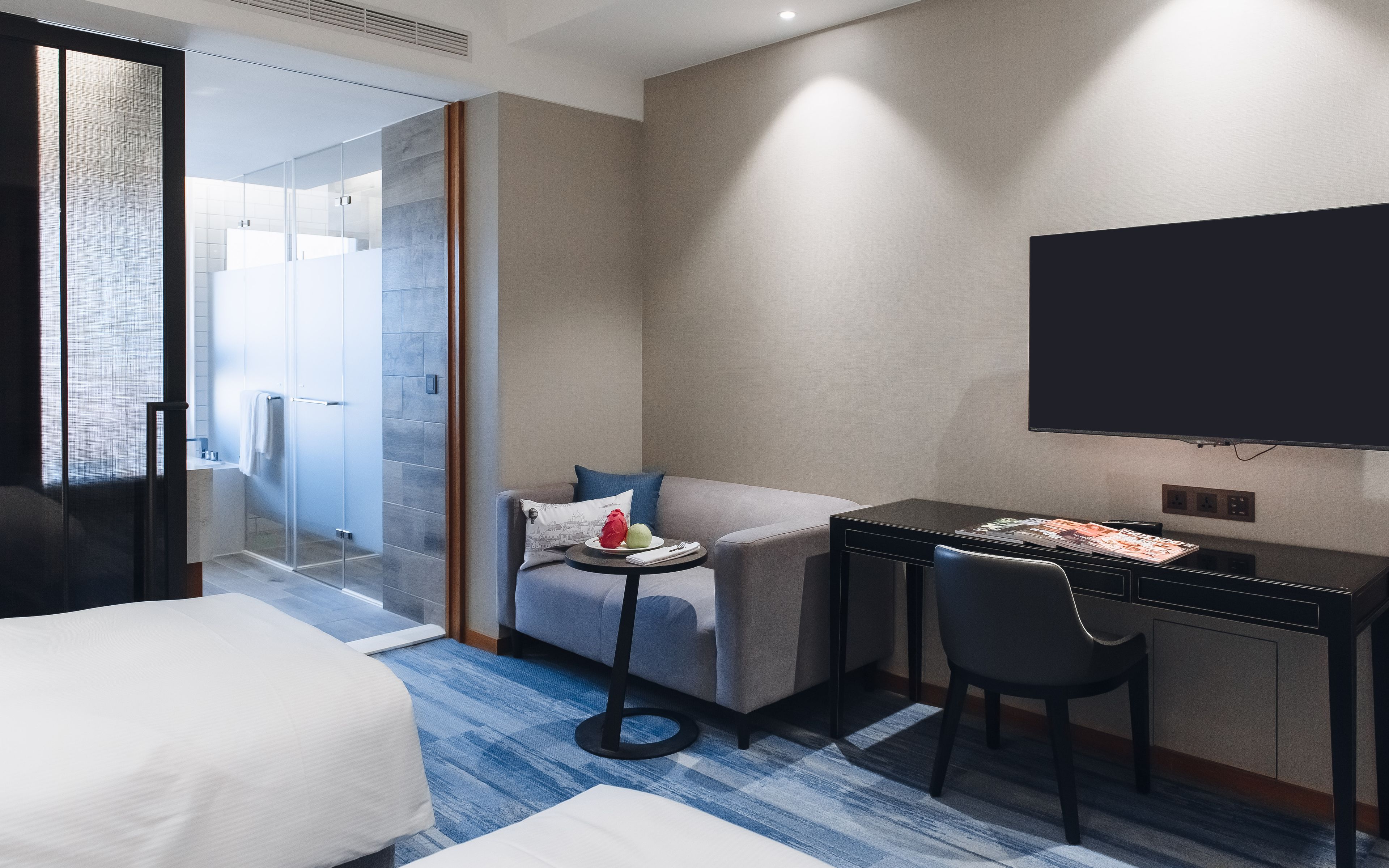 A modern hotel room with a bed, a sofa, a desk with a chair, a television, and a glass shower area.