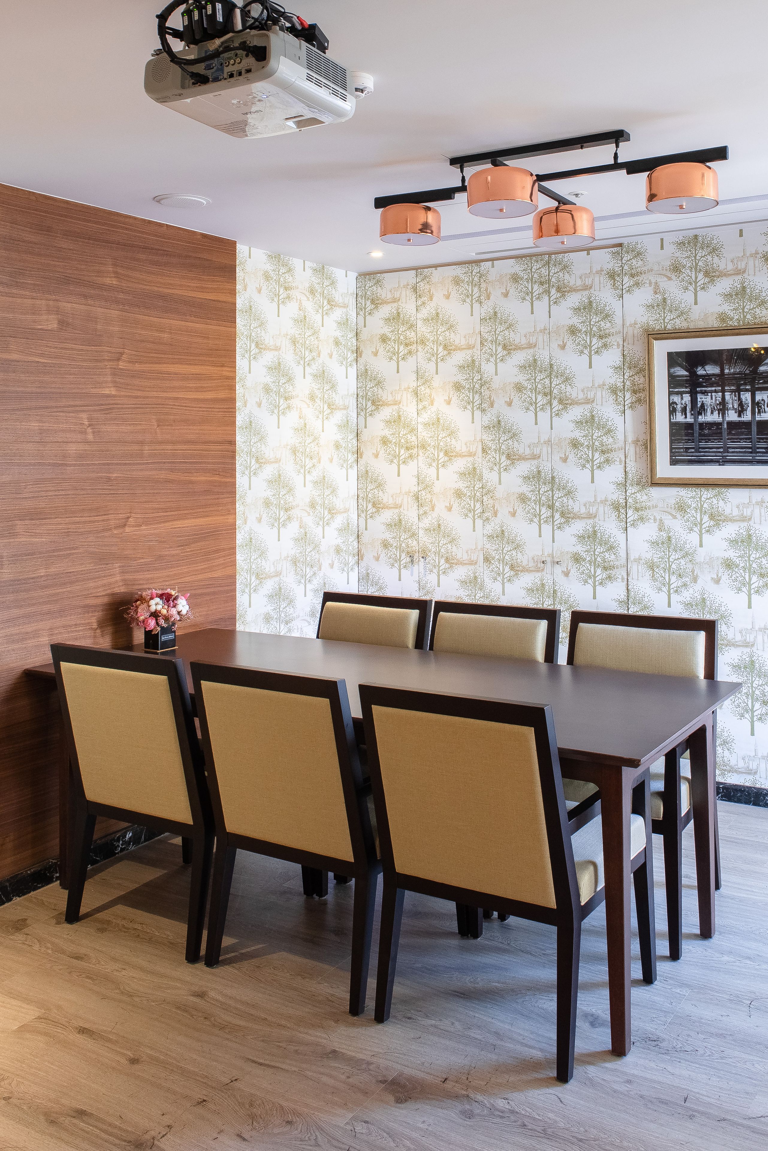 A dining area with a wooden table and chairs, a projector on the ceiling, and a patterned wallpaper with trees.