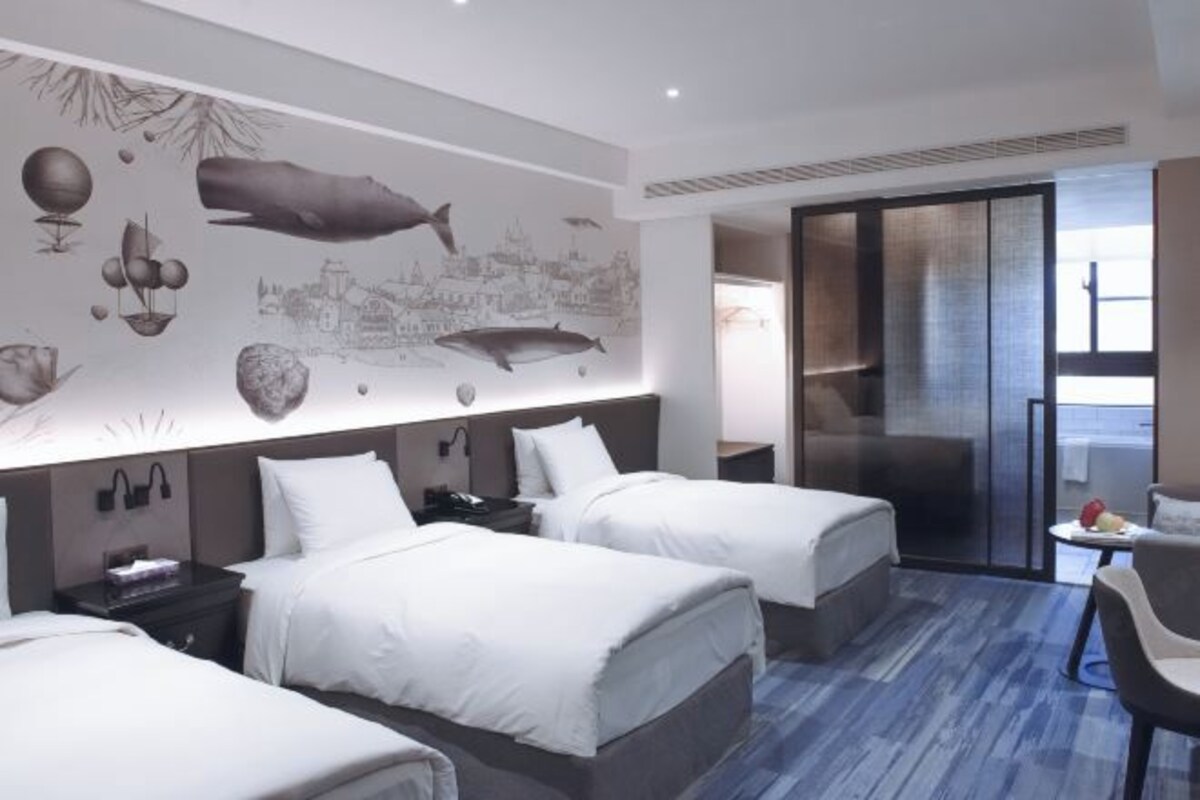 A hotel room with two beds, a large mural on the wall, a sliding door to a balcony, and a small table with fruit.