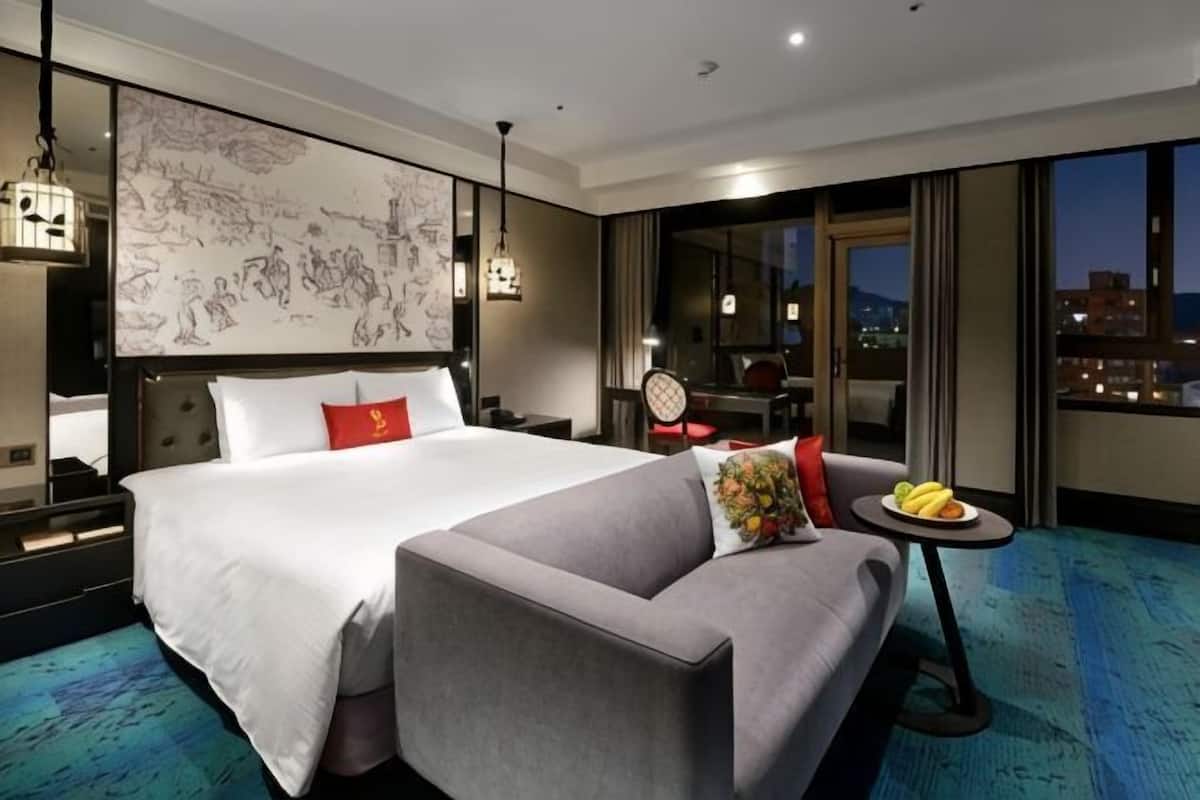 A hotel room with a large bed, a sofa, a small table with fruit, and a city view.