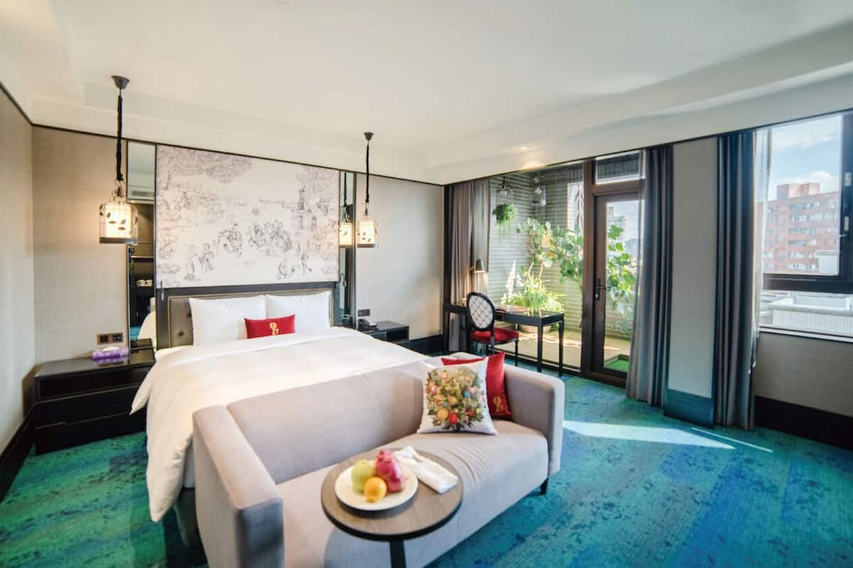 A hotel room with a large bed, a sofa, a small table with fruit, a balcony with plants, and a city view.