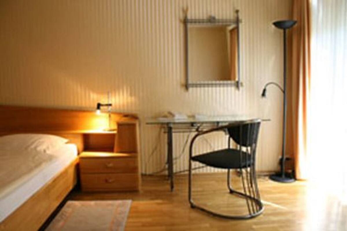 A bedroom with a bed, bedside table, lamp, chair, and a desk with a chair.