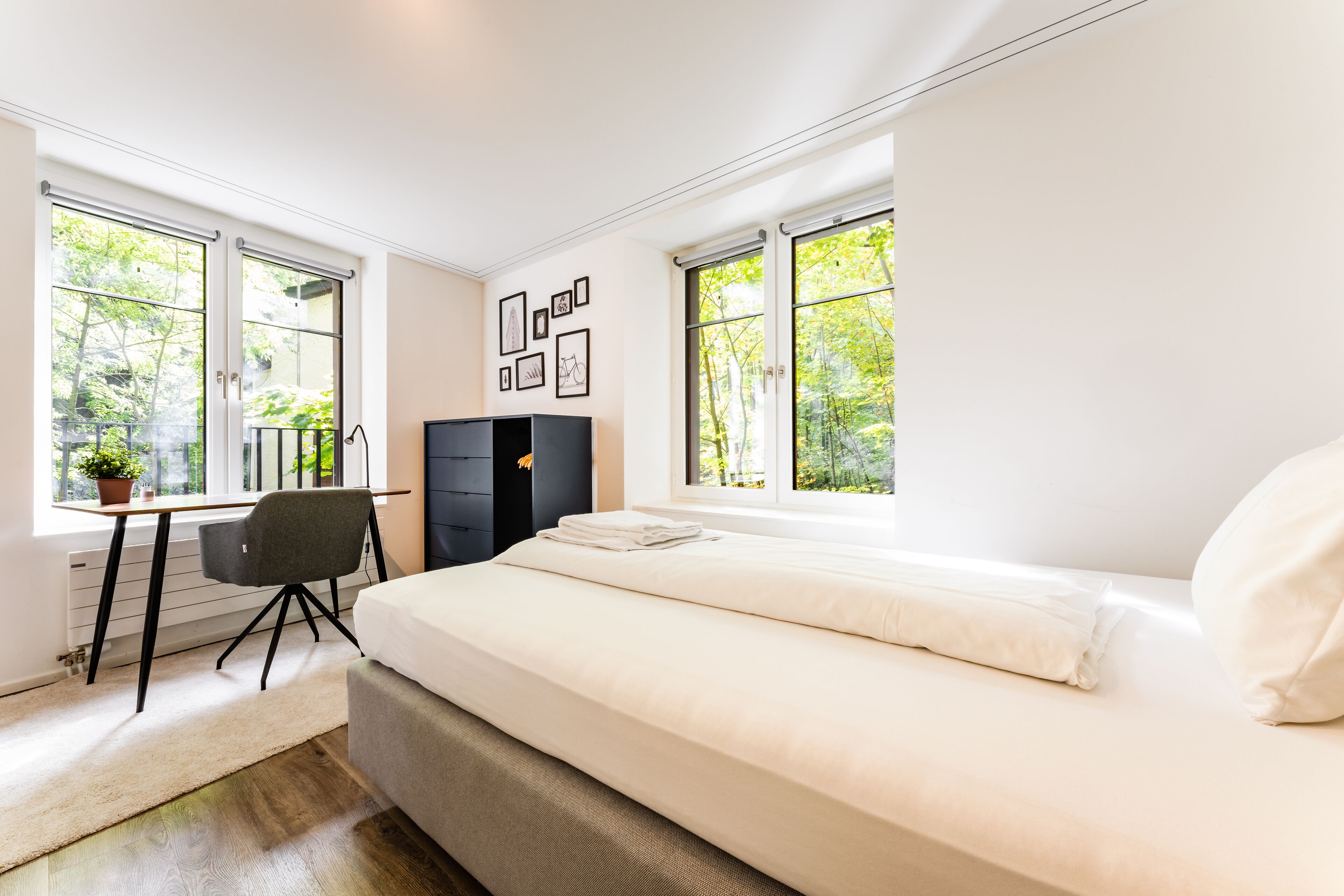 A modern bedroom with a large bed, a desk, and a chair. There are windows with a view of greenery outside.