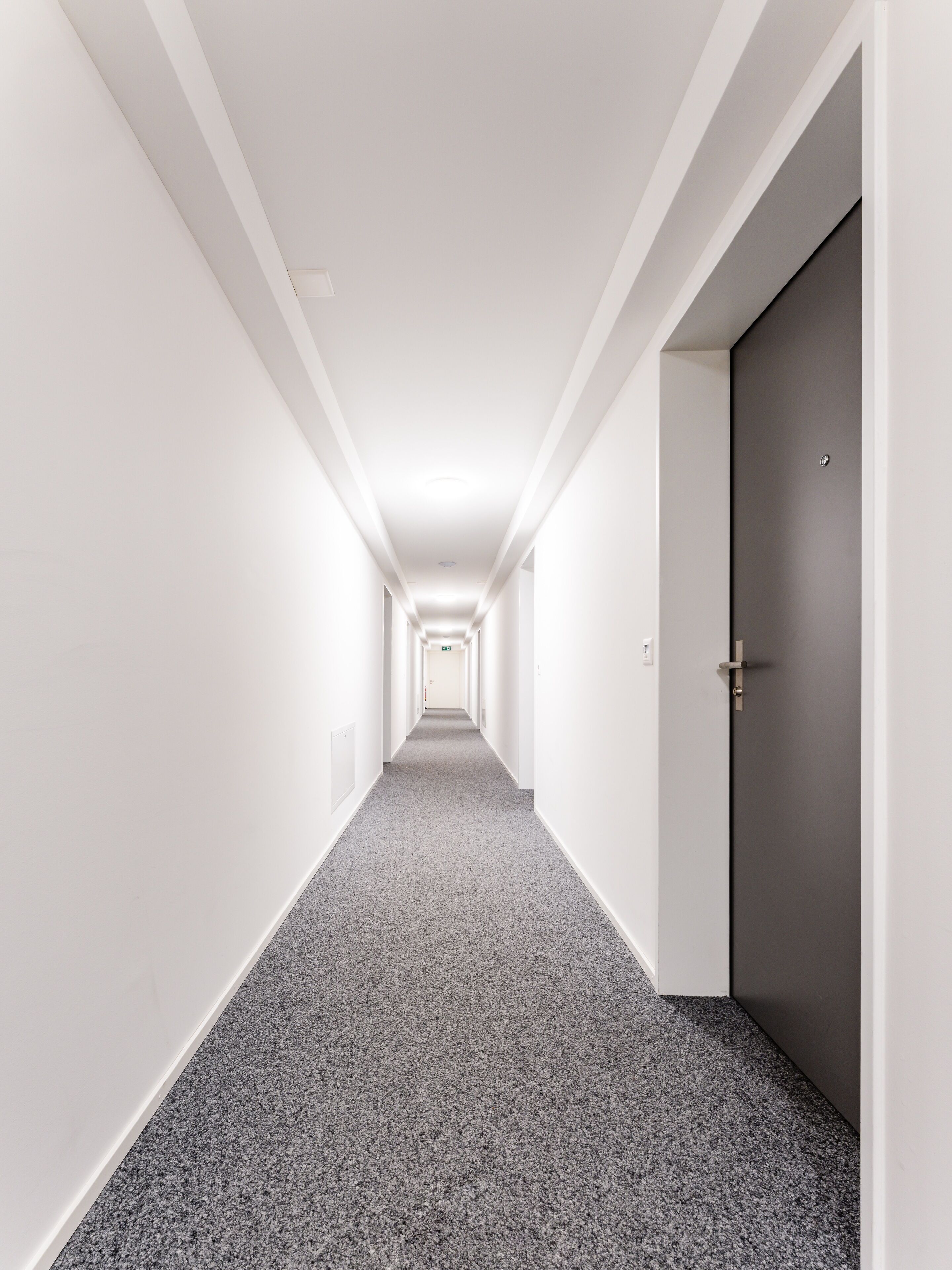 Un long couloir étroit aux murs blancs et au tapis gris.