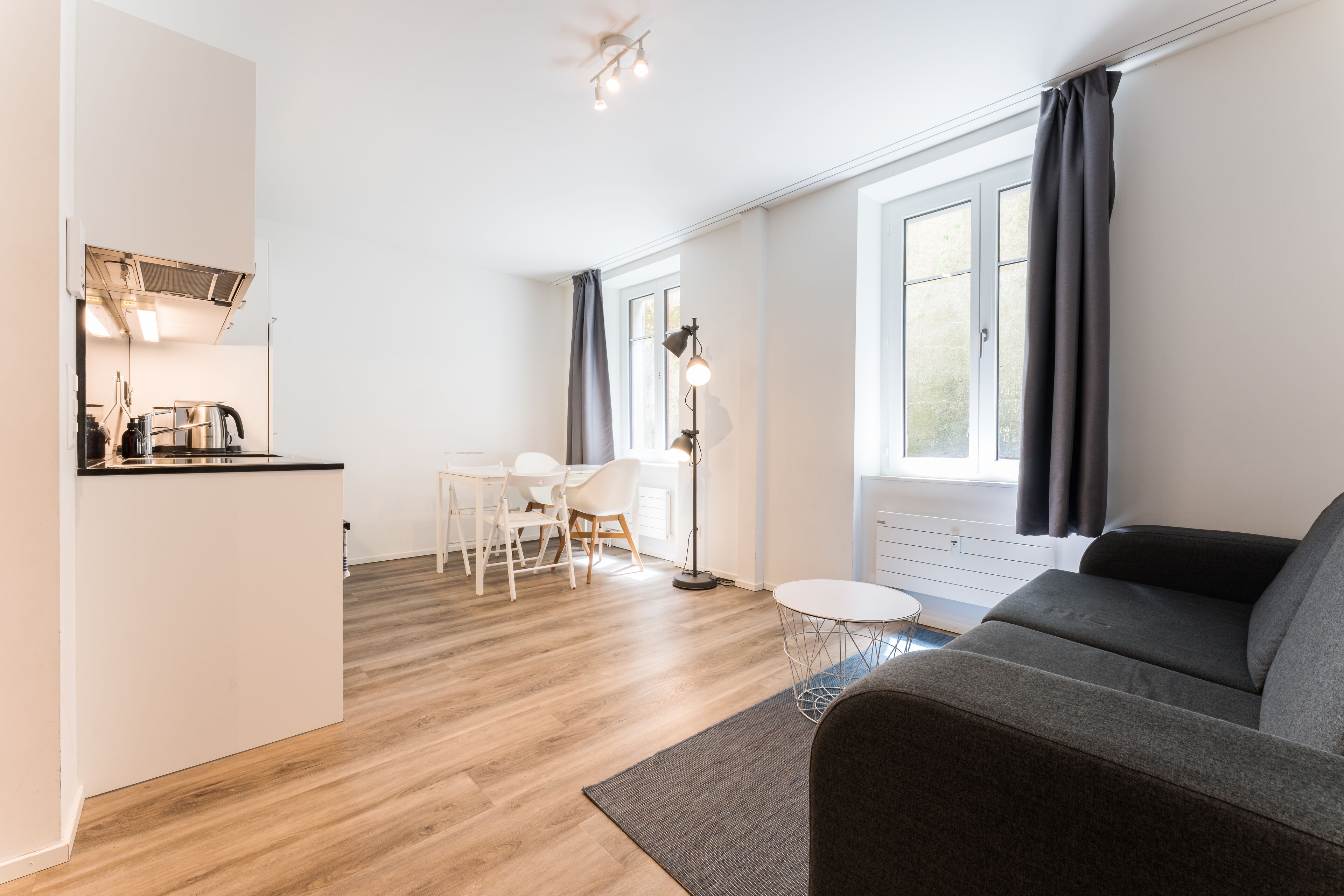 A modern living room with a sofa, a dining table with chairs, a small kitchen area, and a large window with curtains.