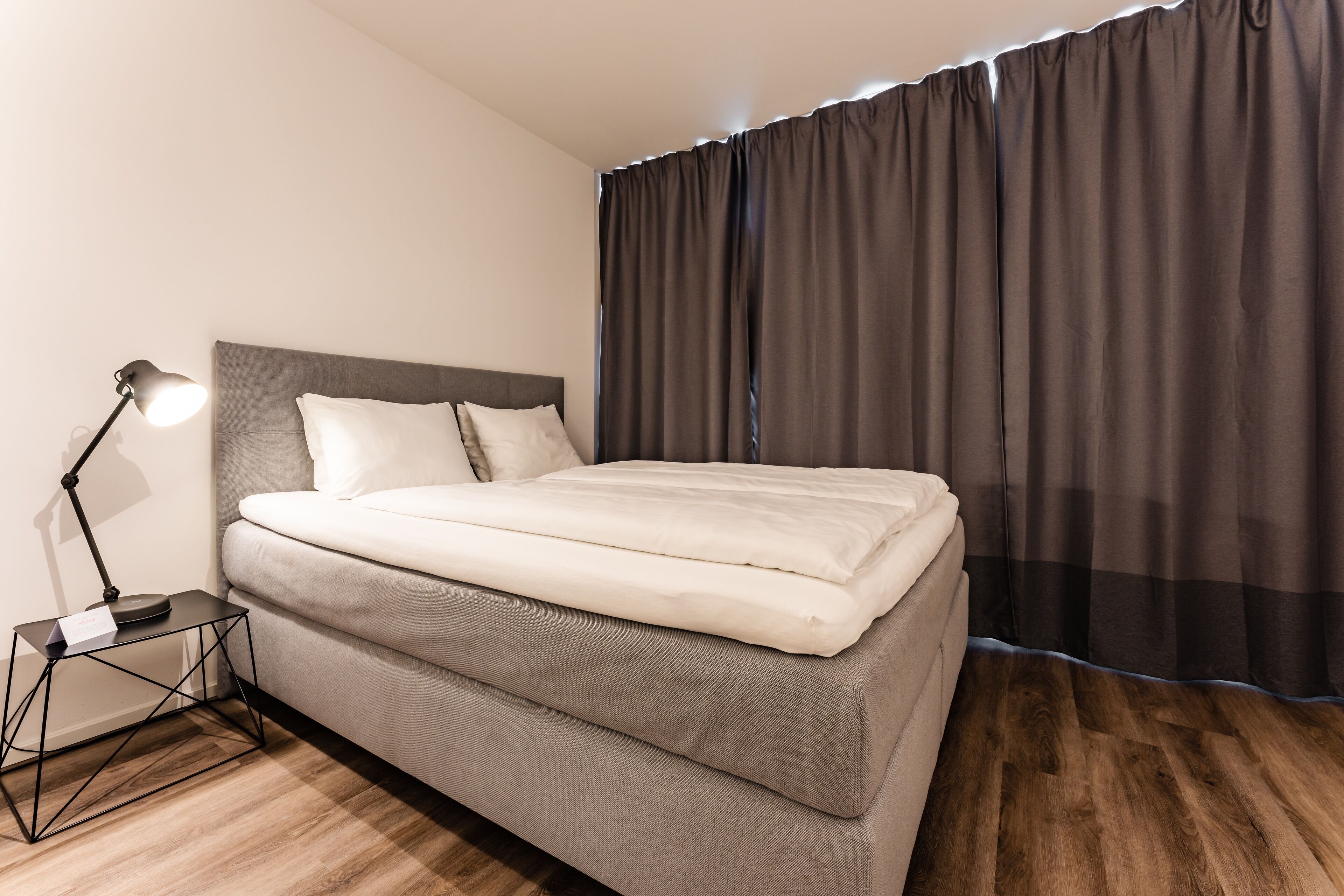 A neatly made bed with a headboard, a bedside table with a lamp, and a window with curtains.