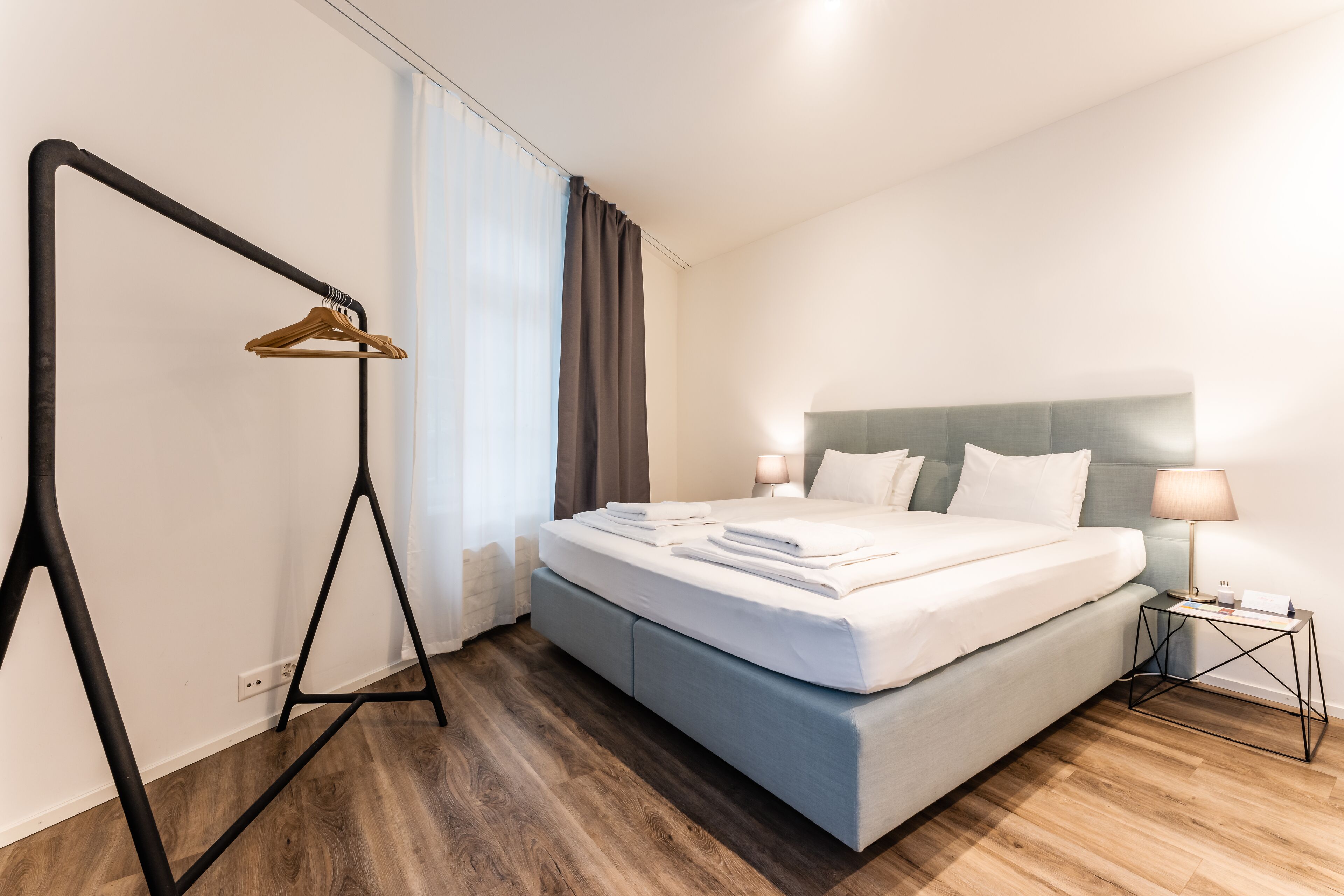 A neatly made bed with white linens, a bedside table with a lamp, and a wooden floor.