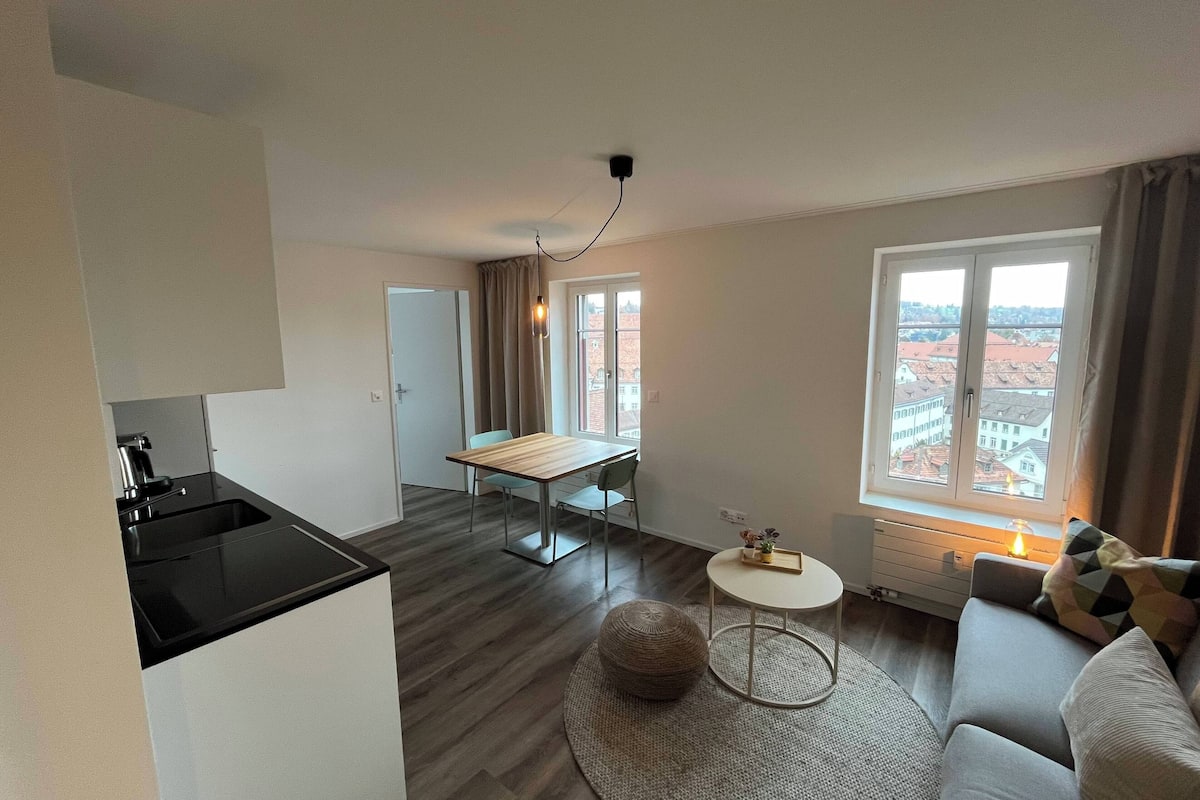 Un apartament modern amb cuina, zona de menjador i saló. Disposa d'un gran finestral amb vista a la ciutat, un otoman rodó i una taula petita rodona.