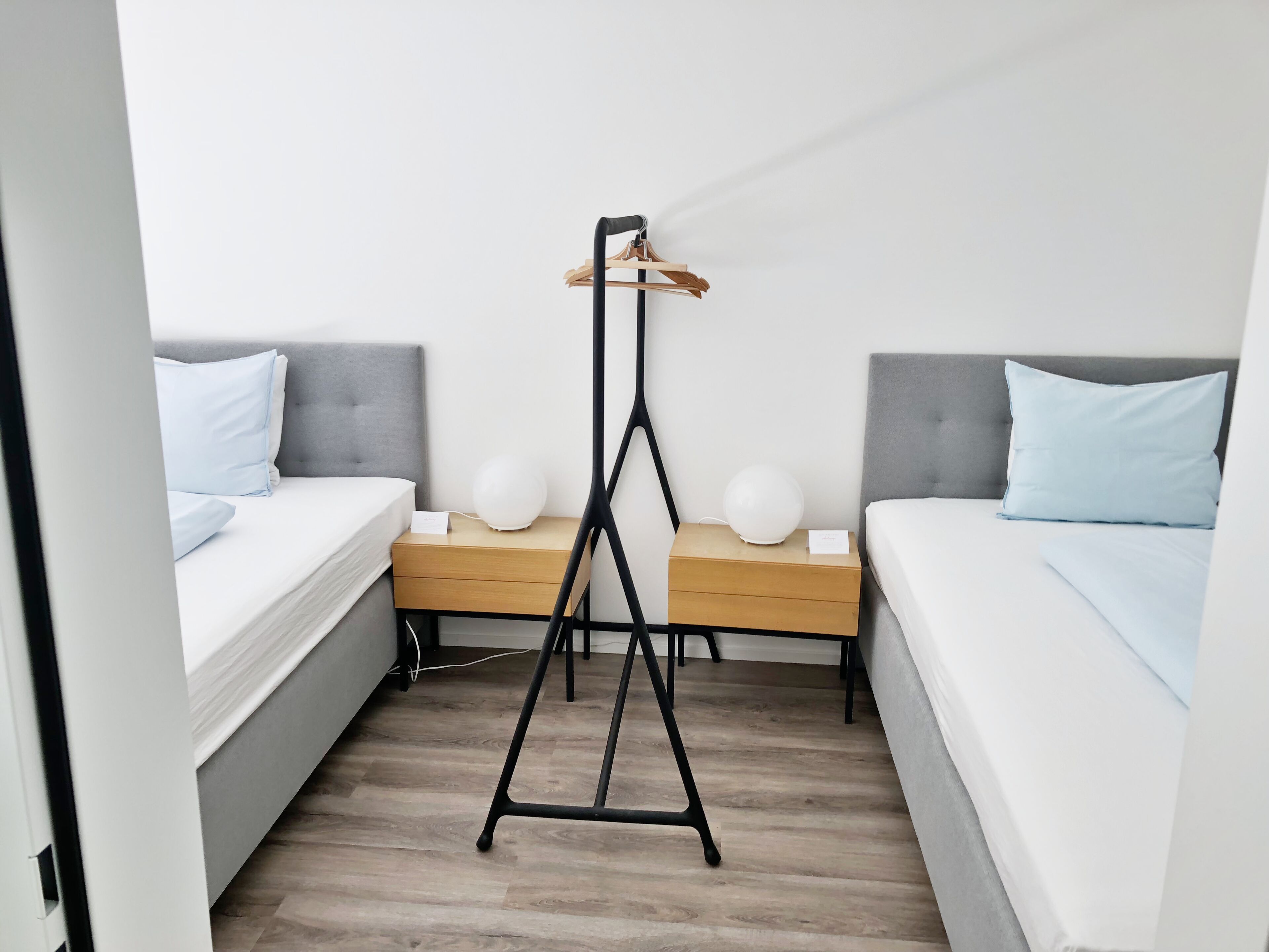 Une chambre d’hôtel avec deux lits, une table de chevet en bois, une lampe de sol noire et un mur blanc.