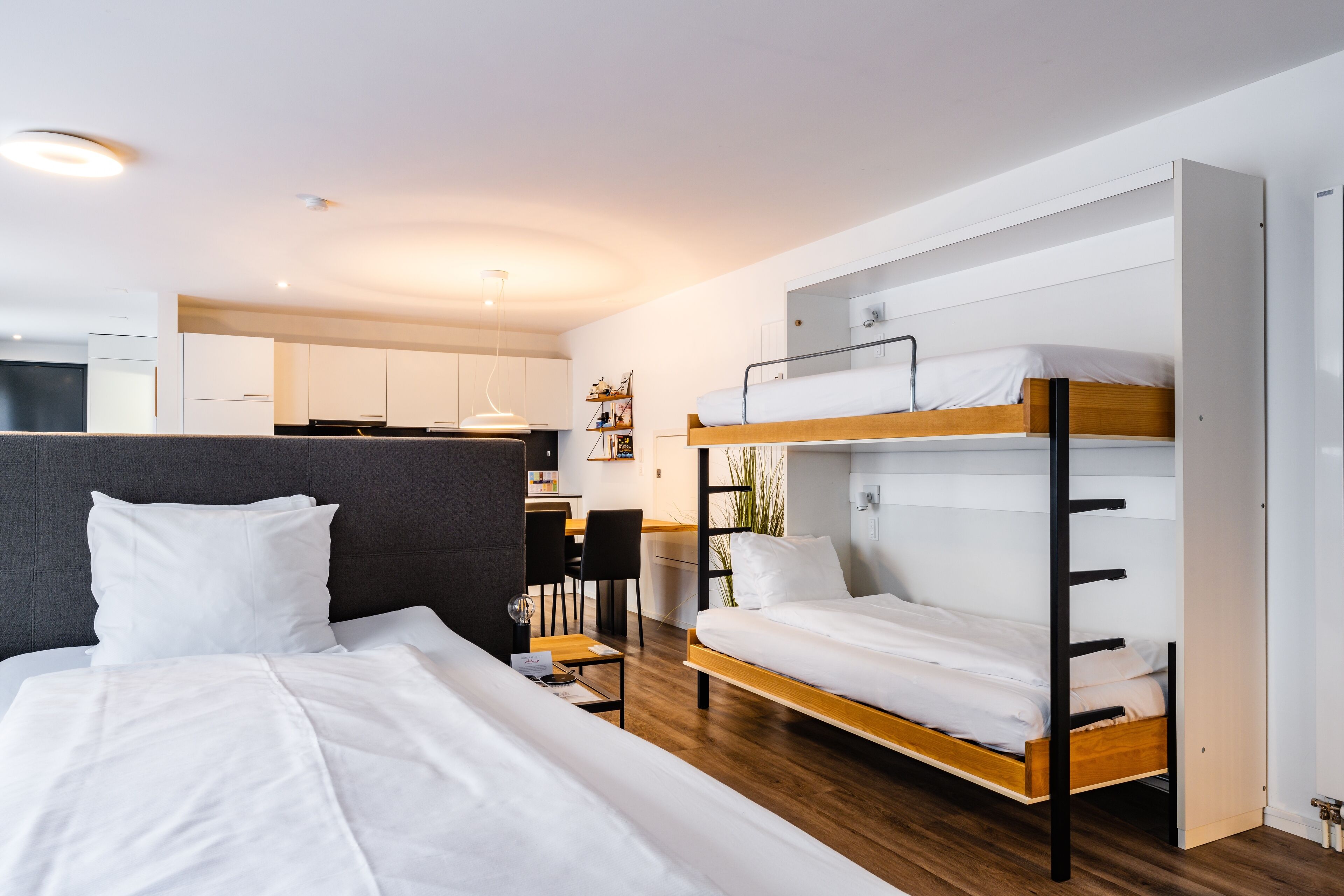Une chambre d’hôtel moderne avec un lit superposé, un lit séparé, un bureau et, au fond, une kitchenette.