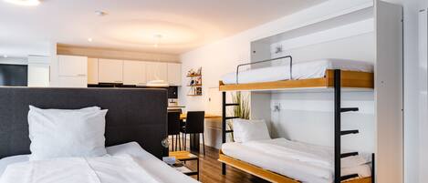 Une chambre d’hôtel moderne avec un lit superposé, un lit séparé, un bureau et, au fond, une kitchenette.
